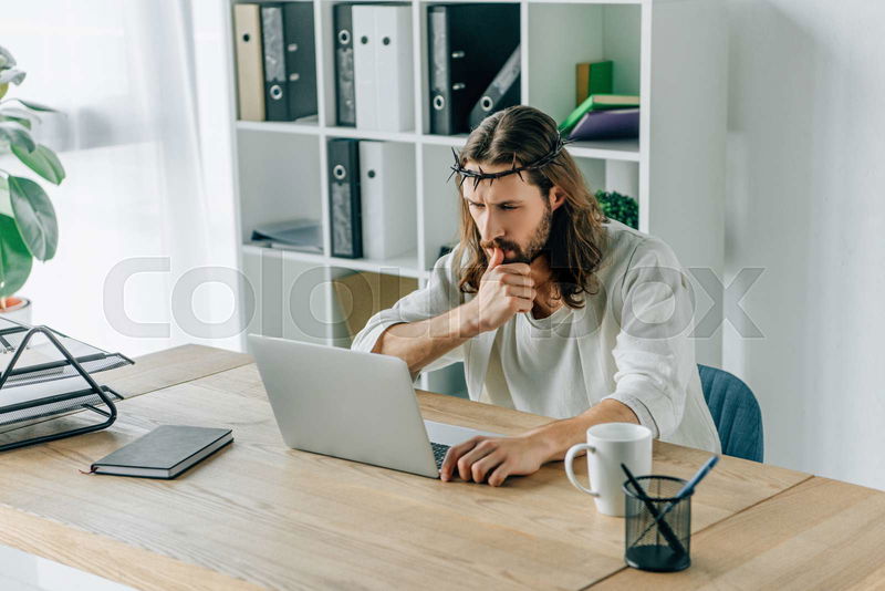 Thoughtful Jesus in crown of thorns using laptop at table in | Colourbox